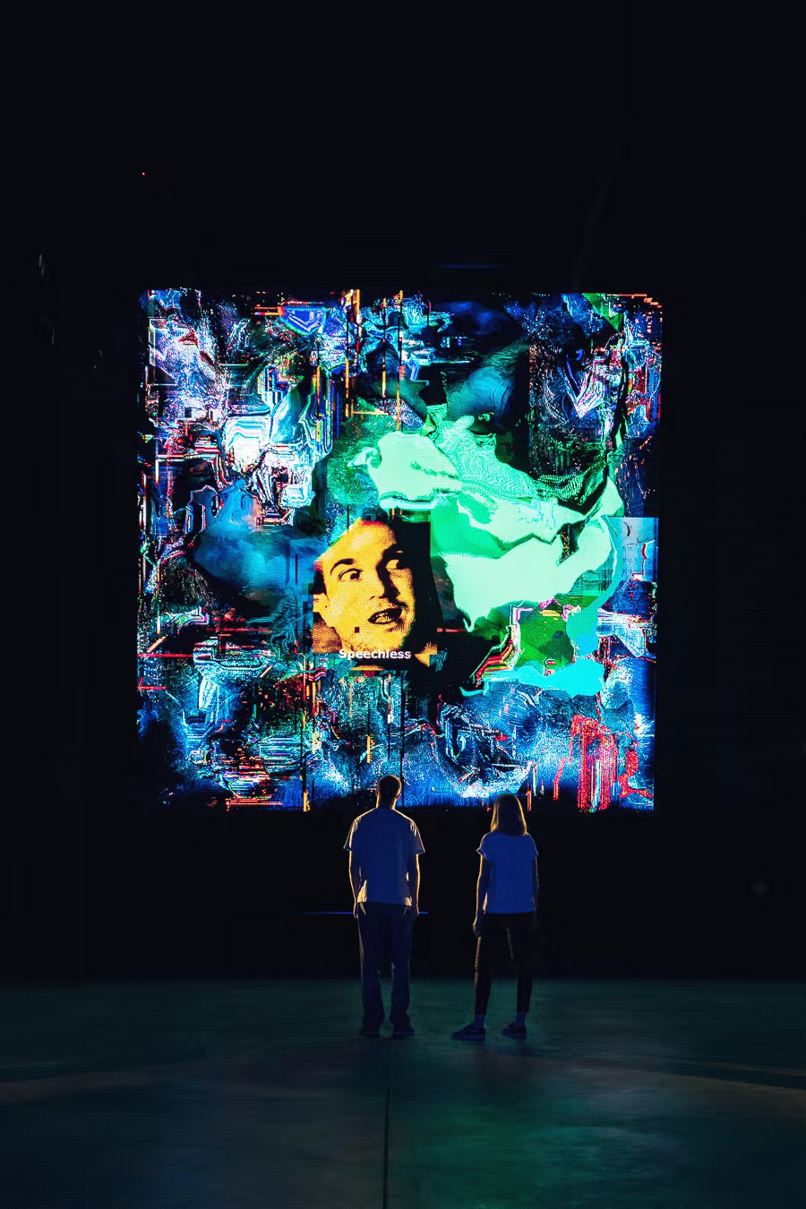 Back views of two people facing large screen with fragmented face and circuit-like overlays in blue tones.