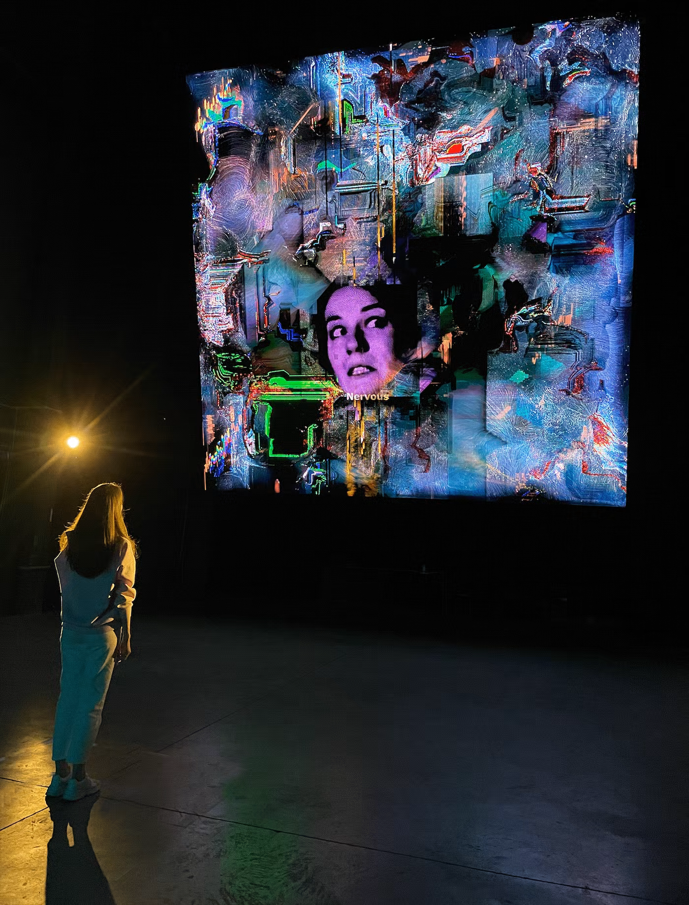 Side view of person in light clothing facing screen with distorted purple face and glowing green overlays.
