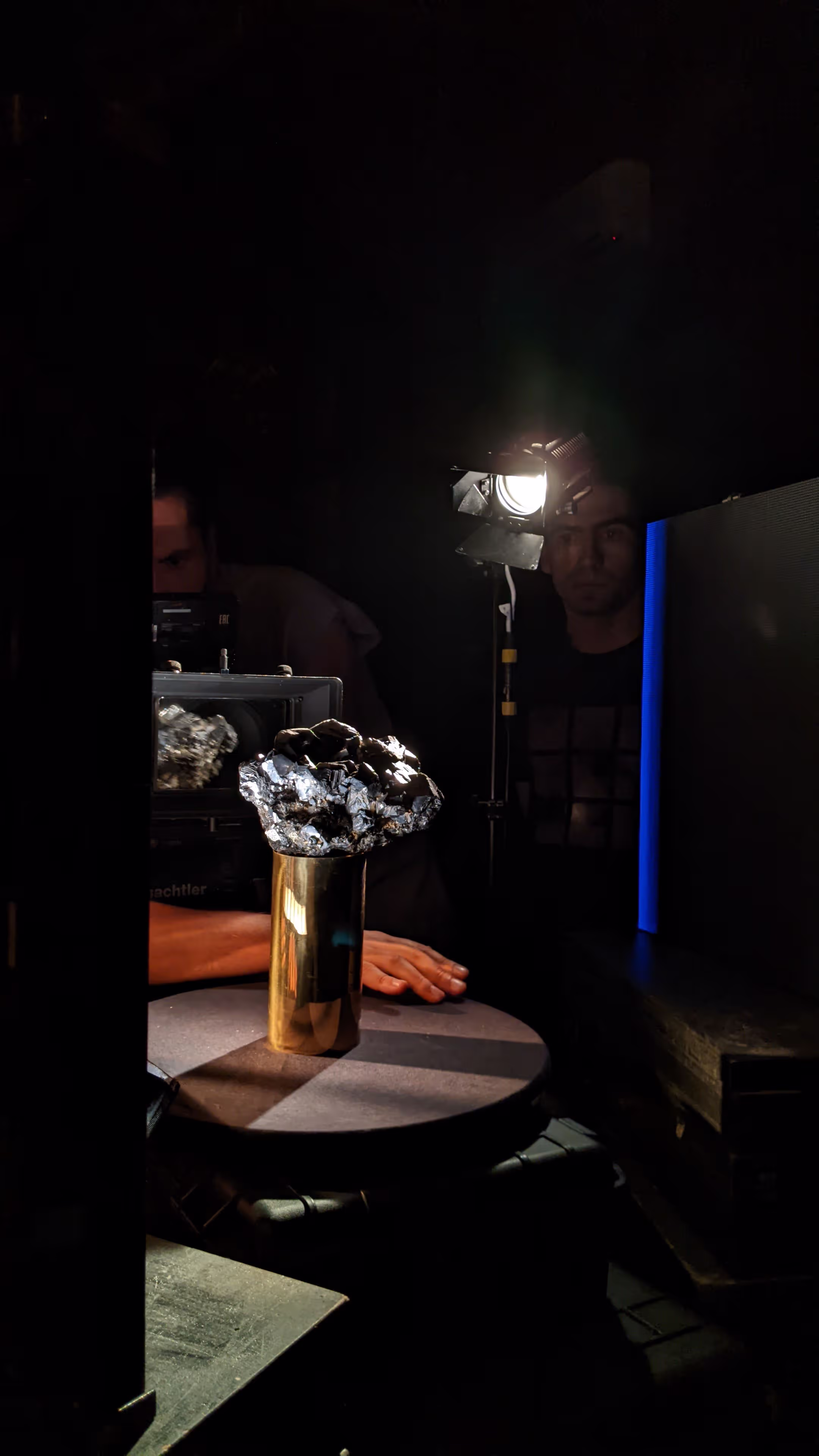 Person's hand near shiny metallic cluster on gold cylinder on table with camera monitor in background.
