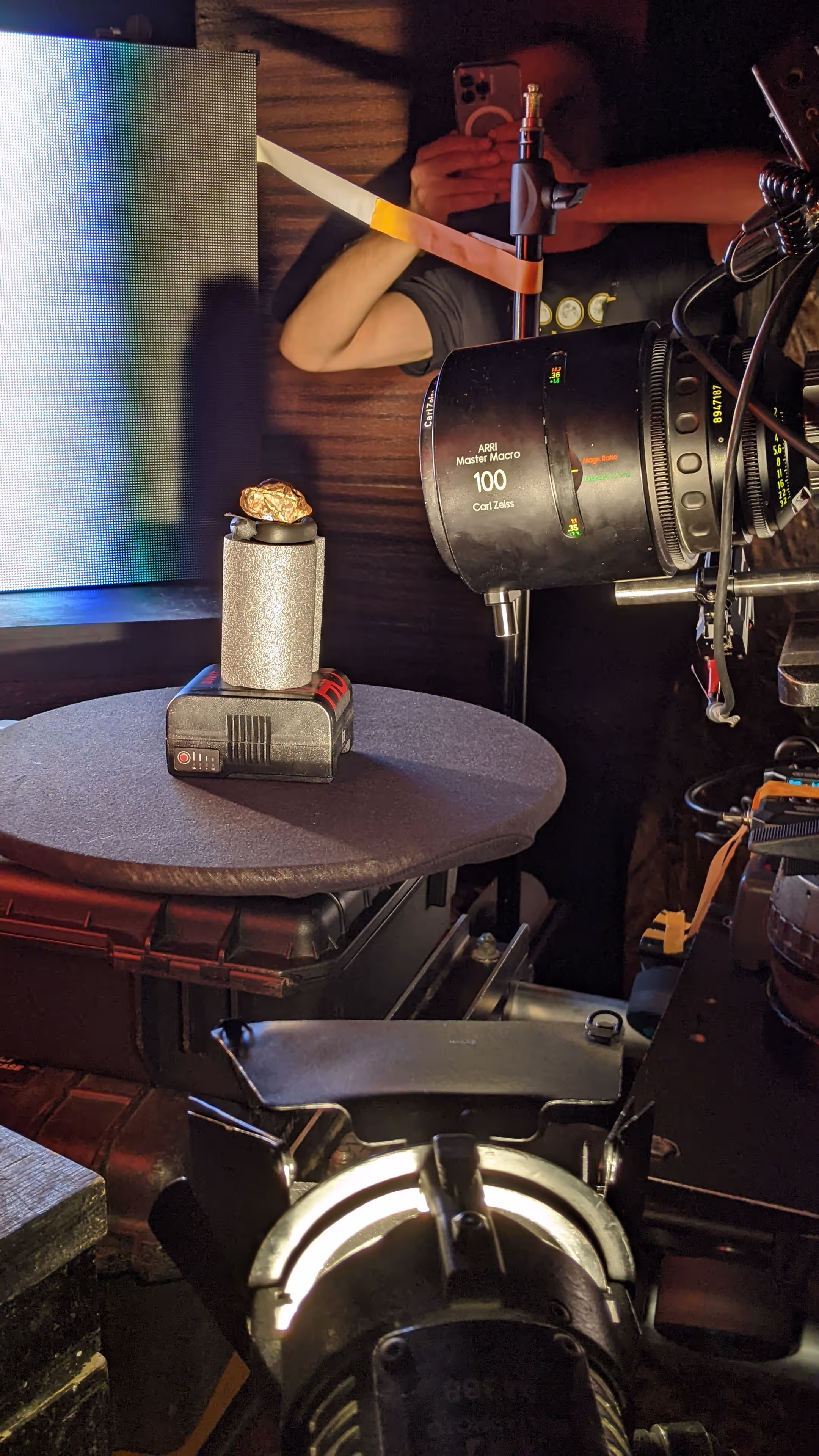 Camera lens over gold ore sample on shiny cylinder base next to black battery pack on table.