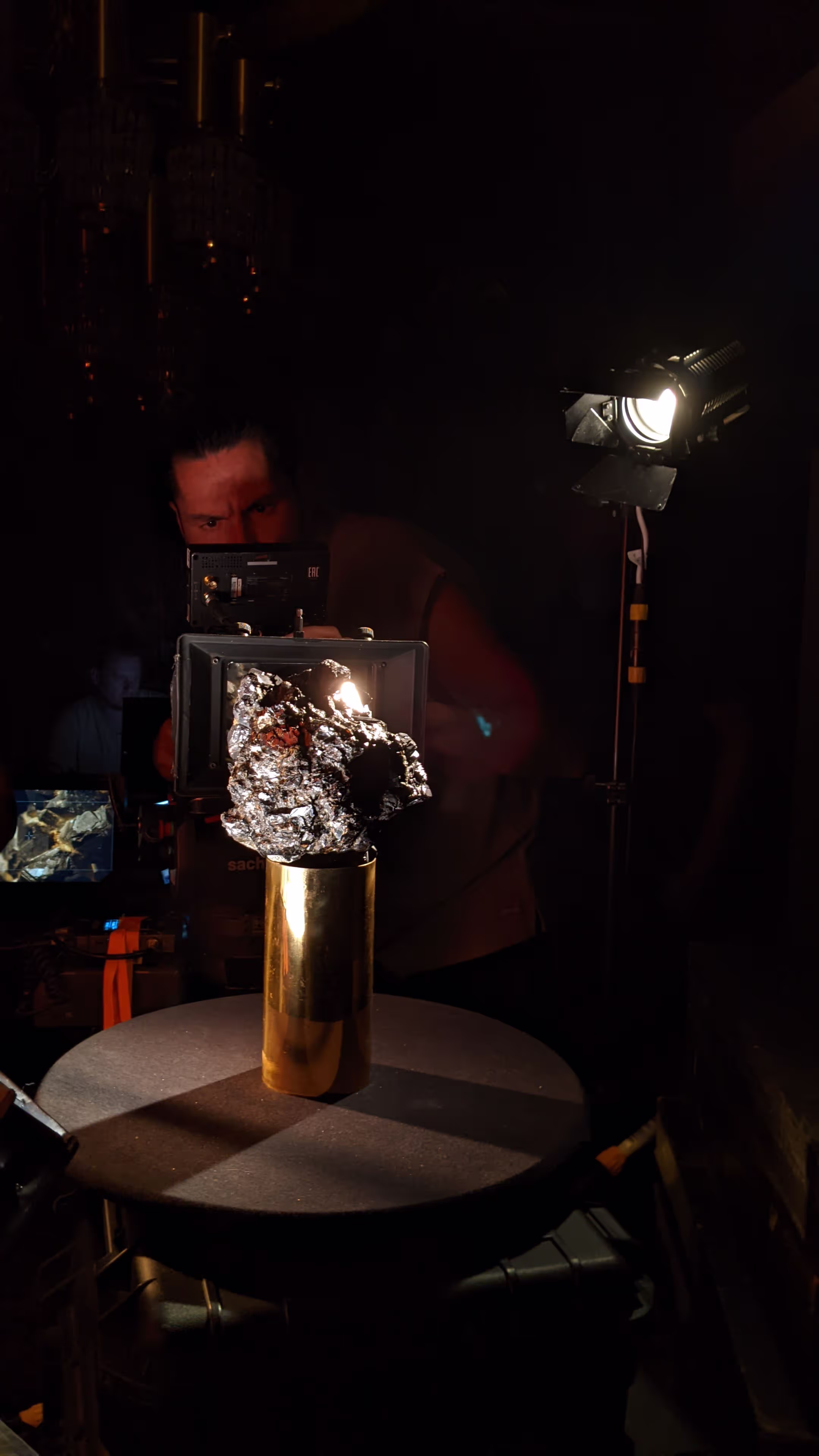 Person operates camera aimed at shiny metallic rock on gold cylinder on round table with lighting equipment.
