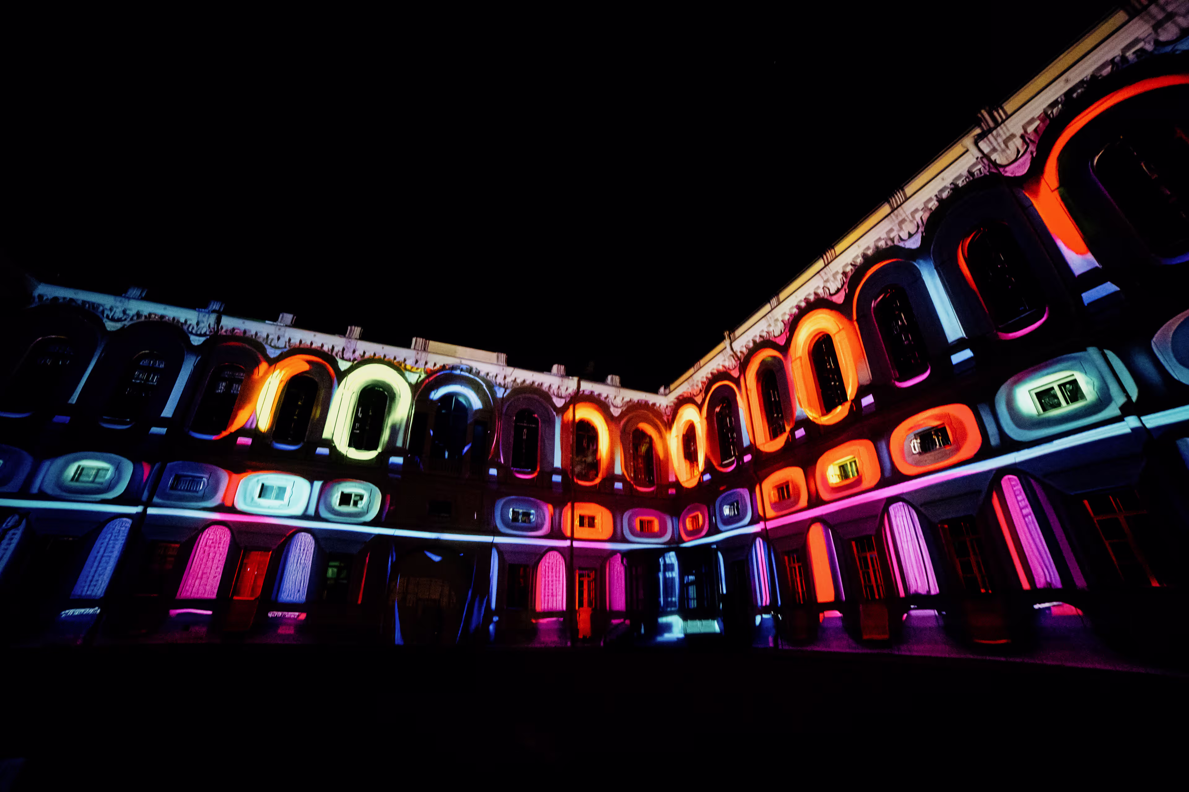 Corner of palace facade with orange, pink, cyan, and magenta glowing arches and windows against dark sky.