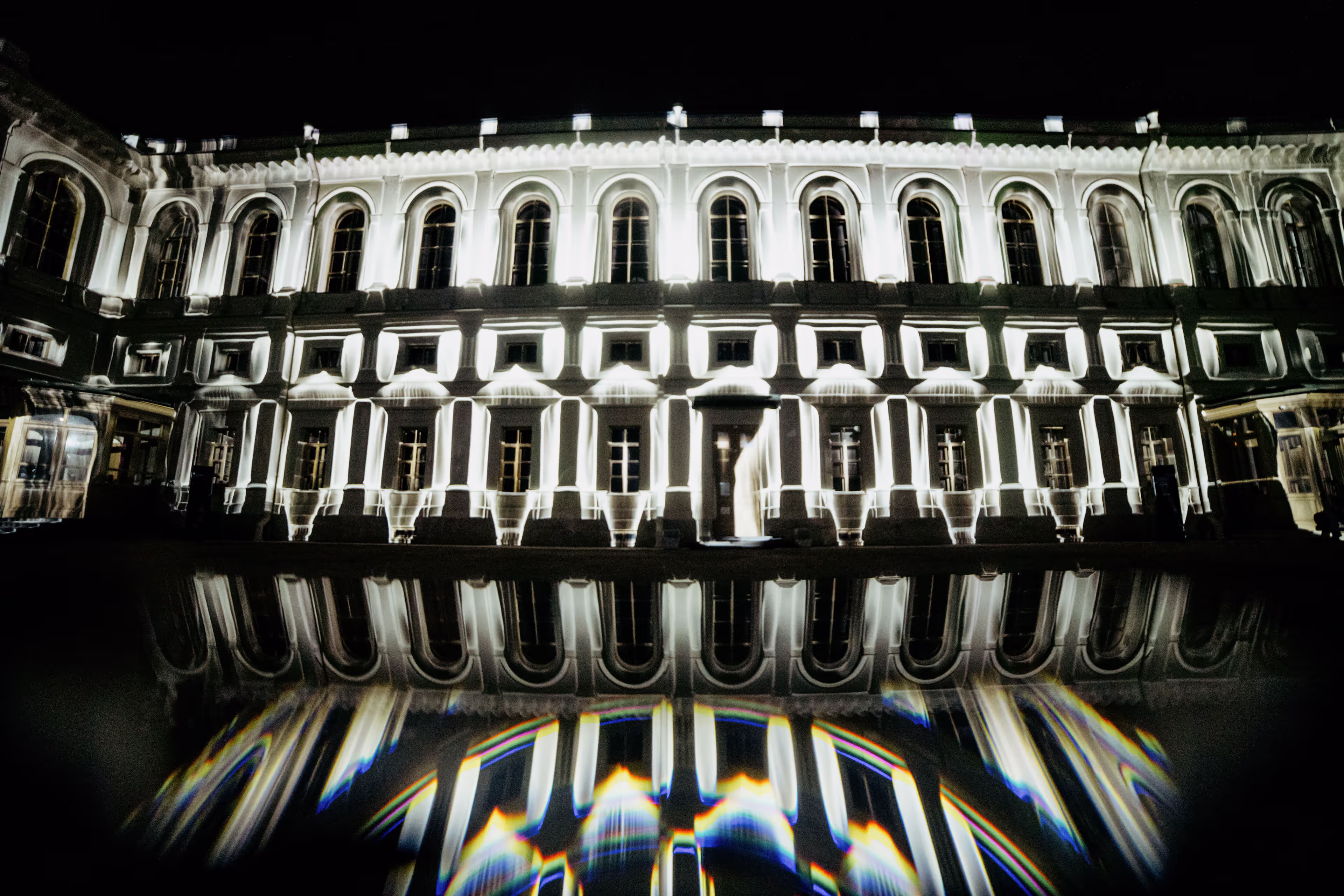 Straight-on view of palace facade with white light projections and prismatic reflections in foreground water.