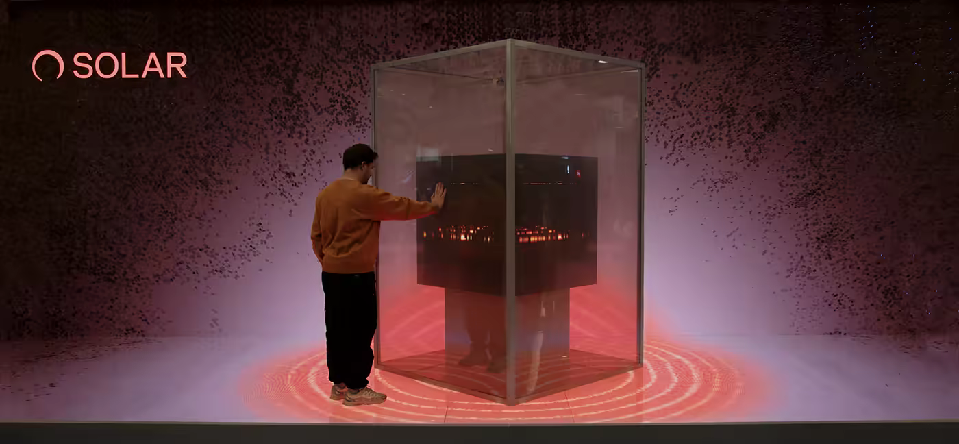 Person in orange sweater hand on glass cube showing red lights inside with red ring on floor.